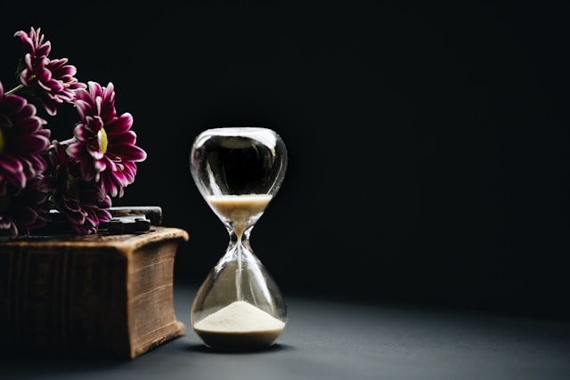 A still life composition symbolizing the refinement of time, featuring a glass hourglass with sand beside a vintage book and purple flowers on a dark background