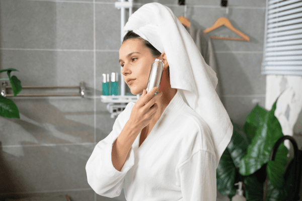A woman in a robe and towel wrap is using a small, handheld electronic device on her cheek as part of a personal beauty routine.