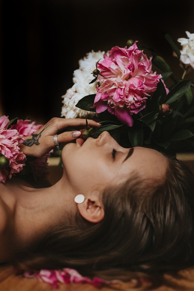 An artistic portrait embodying authentic femininity, showing a woman with eyes closed, gracefully resting among vibrant pink and white peonies